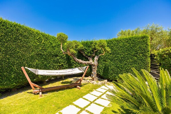 A hammock in a garden with a tree, a stone pathway, and a well-maintained hedge.