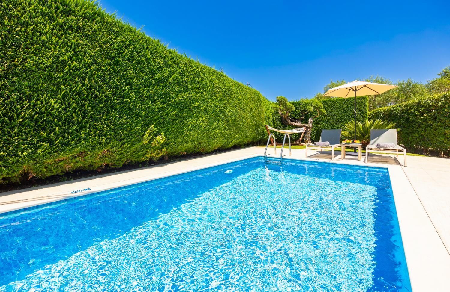 Une piscine rectangulaire à l’eau d’un bleu limpide, entourée d’une terrasse blanche. On y trouve des chaises longues et une petite table avec un parasol. Un arbuste taillé avec soin borde la piscine.