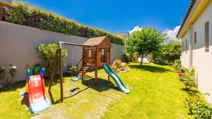 Un parque infantil con un tobogán de madera y columpios, rodeado de un césped cuidado y un edificio blanco.