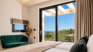 A bedroom with a bed, a green chair, a small table, and a large window with a view of the sea. - Villa Vatia 3 - Three Bedroom Villa, Sleeps 8 (Ionian Islands)