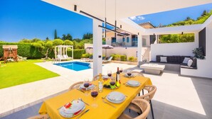 Uma área moderna para refeições ao ar livre com piscina, um espaço coberto para sentar e vista para o verde circundante.