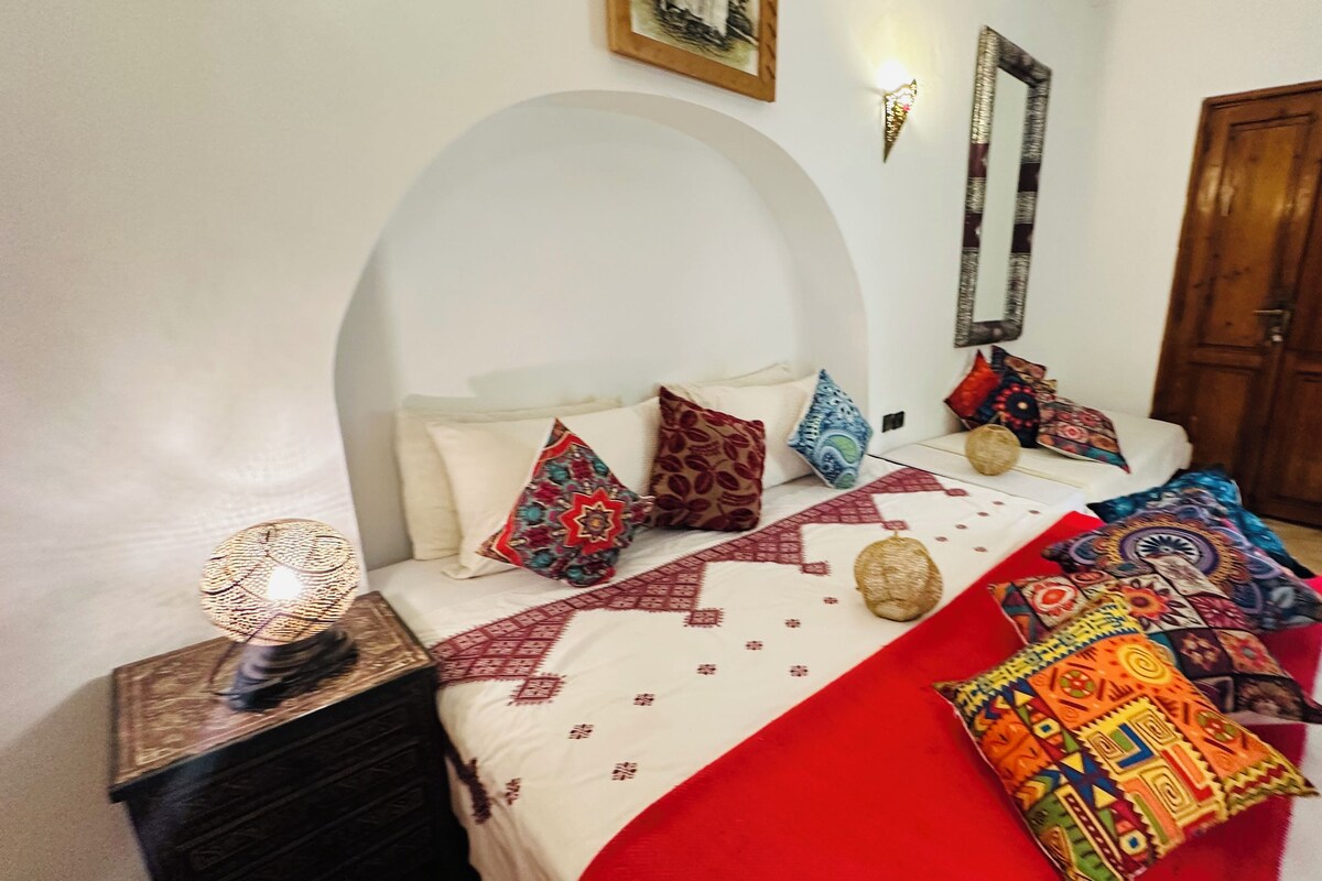 A bed with a red and white bedspread, multiple pillows, a woven basket, and a wooden nightstand with a lamp.