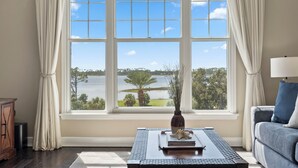Ein Zimmer mit einem großen Fenster, das Blick auf einen See und Bäume bietet. Es gibt einen Tisch mit einer Vase und Büchern, sowie eine Couch mit einer Lampe.