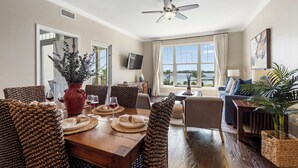Ein geräumiges Wohnzimmer mit großem Fenster und Blick ins Freie, ein Essbereich mit Holztisch, gedeckt für eine Mahlzeit, und einem Deckenventilator.