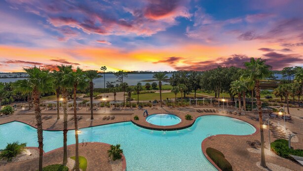 Ein Pool mit geschwungenem Design, umgeben von Palmen und Liegestühlen, vor einem Sonnenuntergangshimmel.