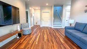 A living room with a wooden floor, a blue sofa, a TV mounted on the wall, and a staircase in the background. - Pelican Point- New construction- Sleeps 11- Pool (St George Island)
