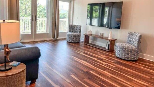 A living room with a wooden floor, a sofa, two patterned armchairs, a TV mounted on the wall, and a balcony visible through the window. - Pelican Point- New construction- Sleeps 11- Pool (St George Island)