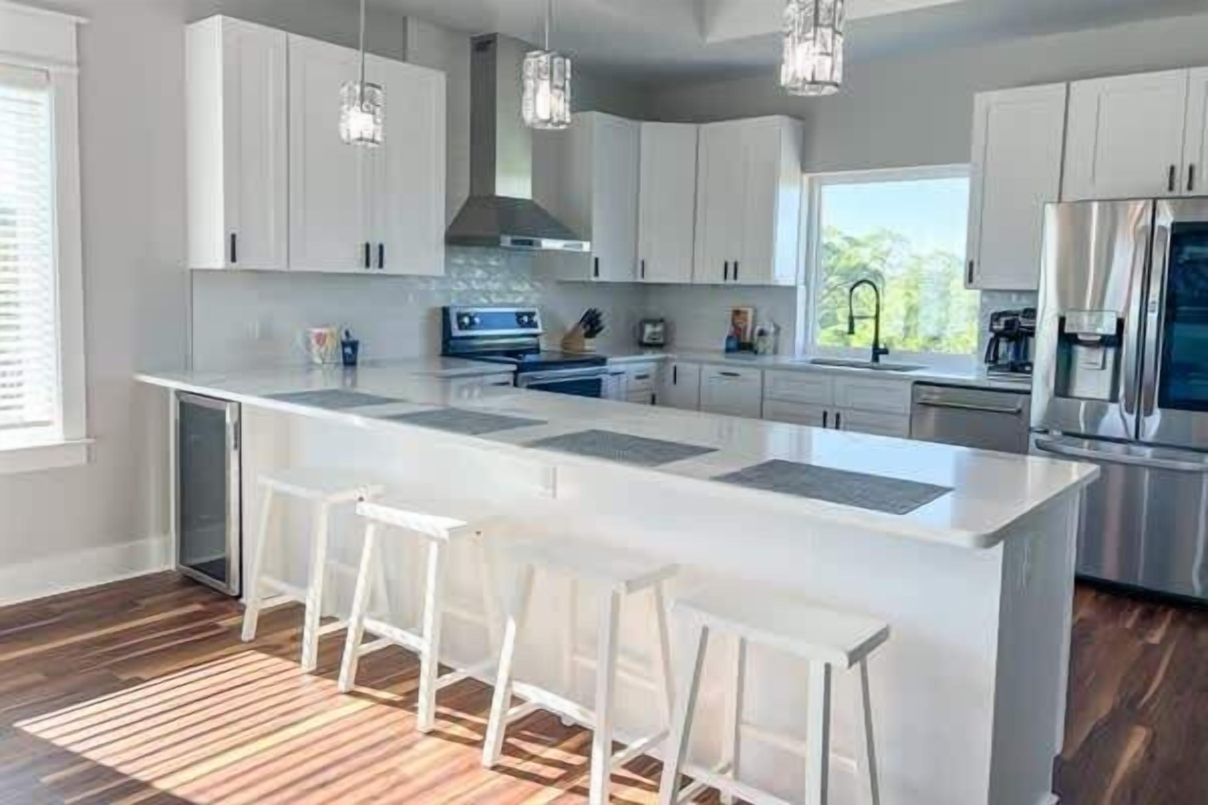 Una cocina moderna con gabinetes blancos, una isla grande con taburetes de bar, electrodomésticos de acero inoxidable e iluminación colgante.