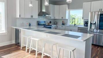 Una cocina moderna con gabinetes blancos, una isla grande con taburetes de bar, electrodomésticos de acero inoxidable e iluminación colgante.