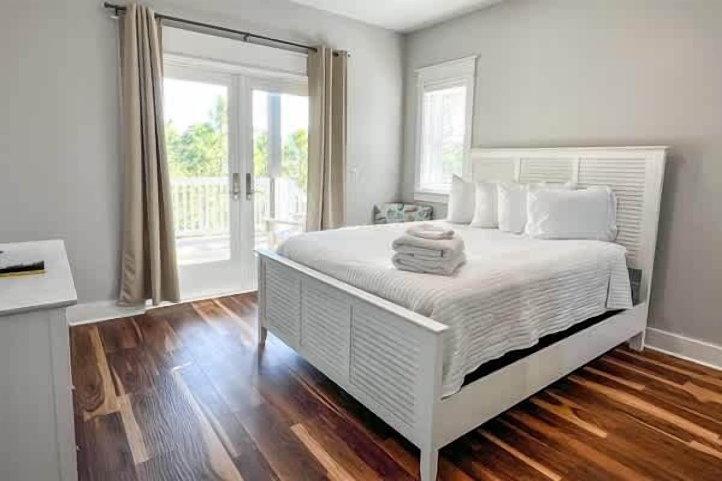 Une chambre avec un lit blanc, un plancher en bois et un balcon agrémenté de rideaux.