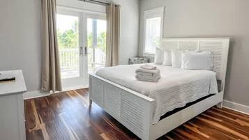 A bedroom with a white bed, wooden flooring, and a balcony with curtains.
