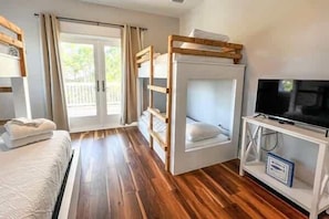 A room with bunk beds, a television on a stand, and a sliding glass door leading to an outdoor area. - Pelican Point- New construction- Sleeps 11- Pool (St George Island)