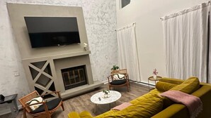 A living room with a yellow sofa, a fireplace, a flat-screen TV mounted on the wall, and a small round table with a plant.