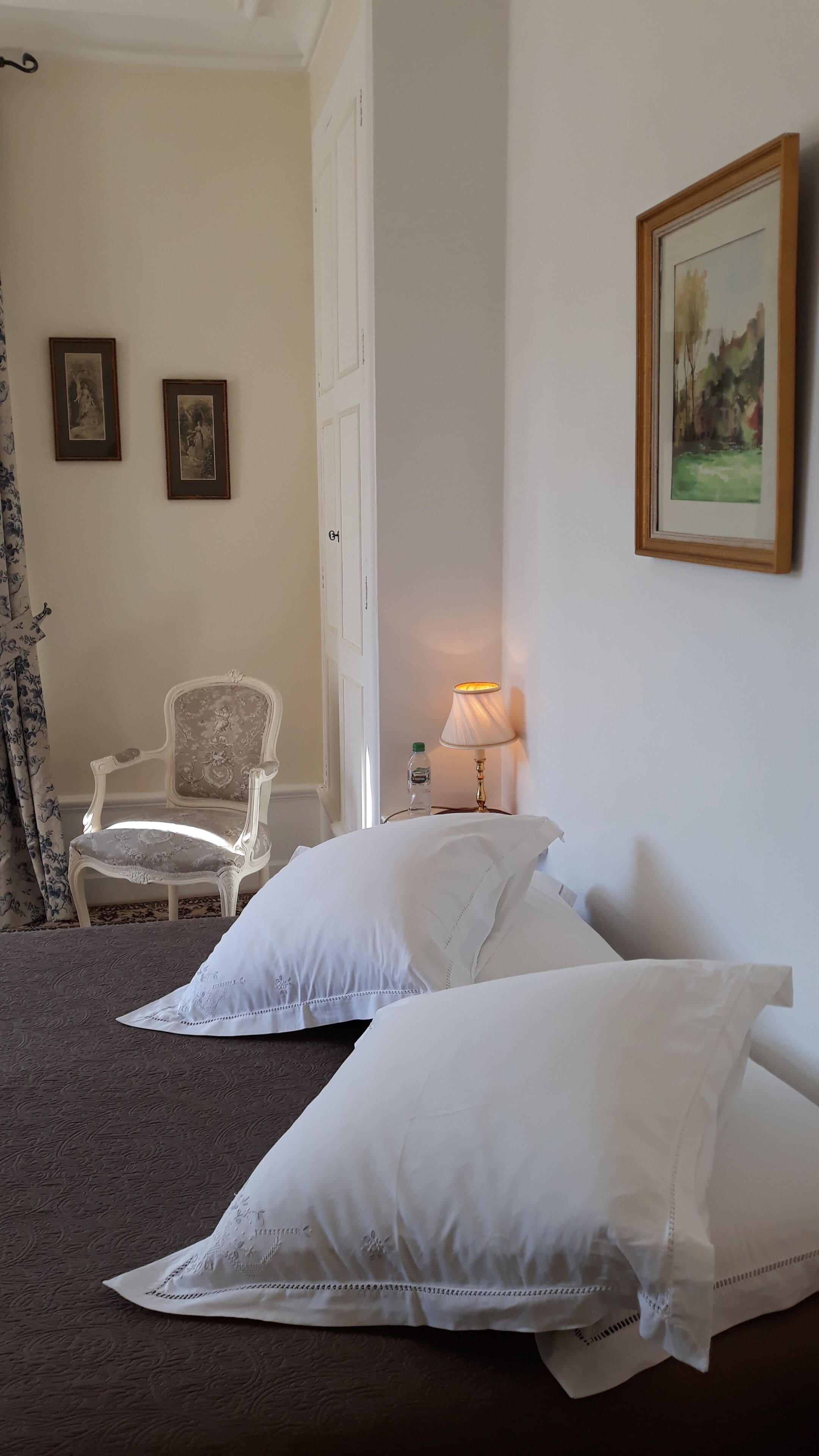 A bedroom with two white pillows, a chair, a bedside table with a lamp, and two framed pictures on the wall.
