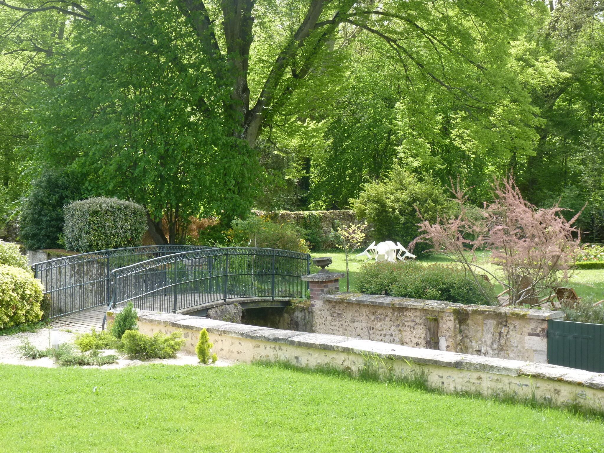 A stone bridge with a metal railing over a small stream, surrounded by lush greenery and trees.