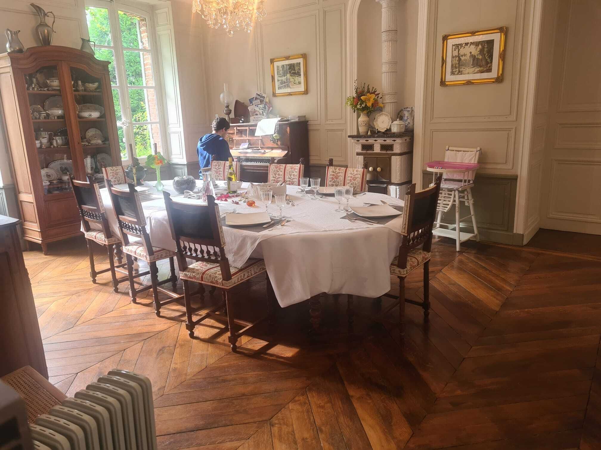Une salle à manger avec une table ronde dressée pour six personnes, entourée de chaises en bois. Un buffet rempli de vaisselle se trouve à gauche, et une cheminée avec une manteau de cheminée est visible au fond.