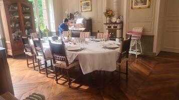 Une salle à manger avec une table ronde dressée pour six personnes, entourée de chaises en bois. Un buffet rempli de vaisselle se trouve à gauche, et une cheminée avec une manteau de cheminée est visible au fond.