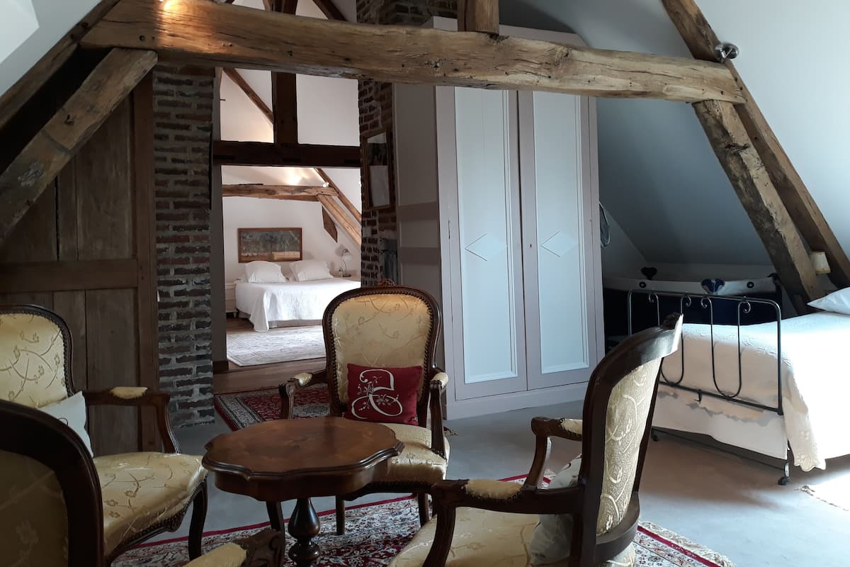Un dormitorio acogedor en el ático con vigas de madera a la vista, una cama, dos sillones y una mesita.