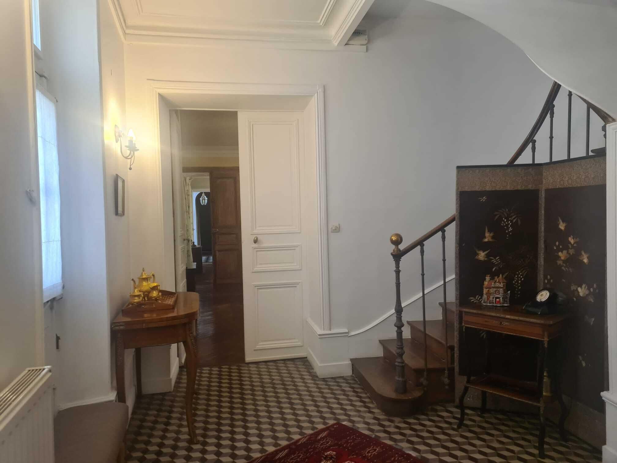 Un couloir doté d’un sol à carreaux, un escalier avec une rampe en bois et une petite table sur laquelle se trouve une lampe.