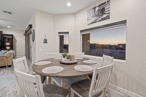 A dining area with a round table, white chairs, and a view through a large window.