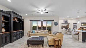 A spacious living area with a large sofa, a coffee table, and a dark-colored ottoman. There is a kitchen area with a dining table and chairs visible in the background.