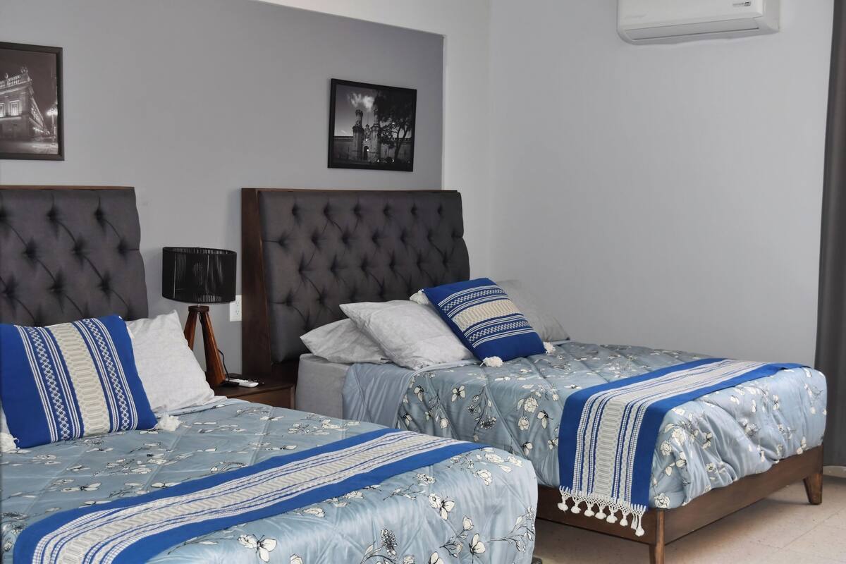 A bedroom with two beds, a wall-mounted air conditioner, and two framed pictures.