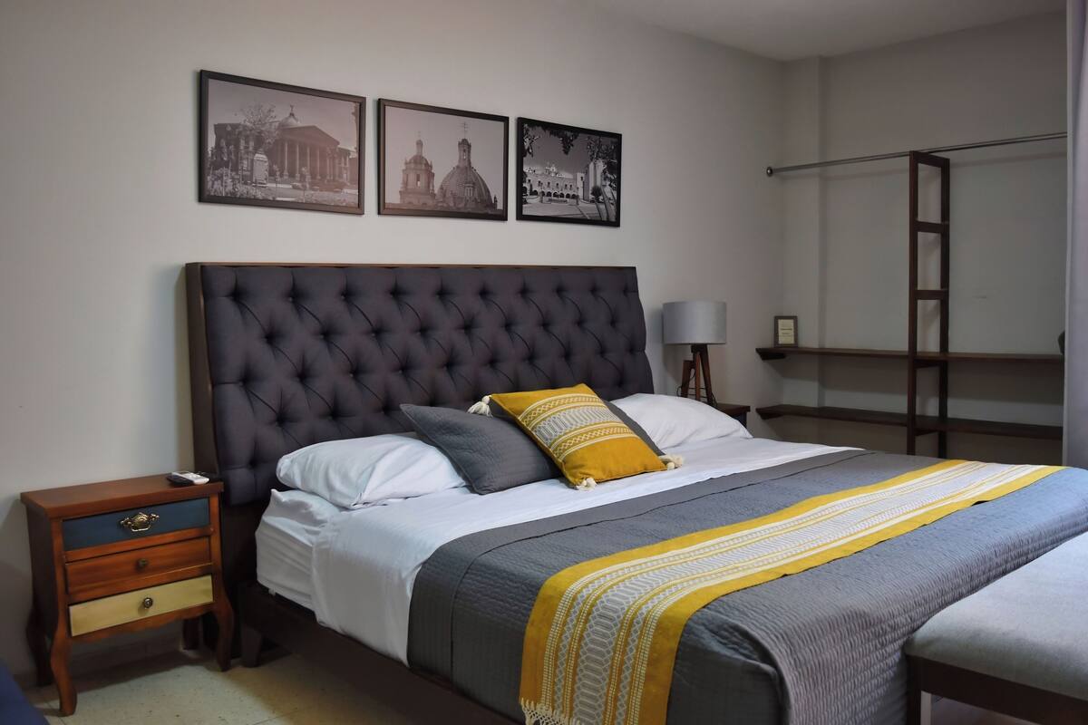 A bedroom with a tufted headboard, a bed with white and gray bedding, a mustard-colored throw, and three black-and-white framed pictures on the wall.