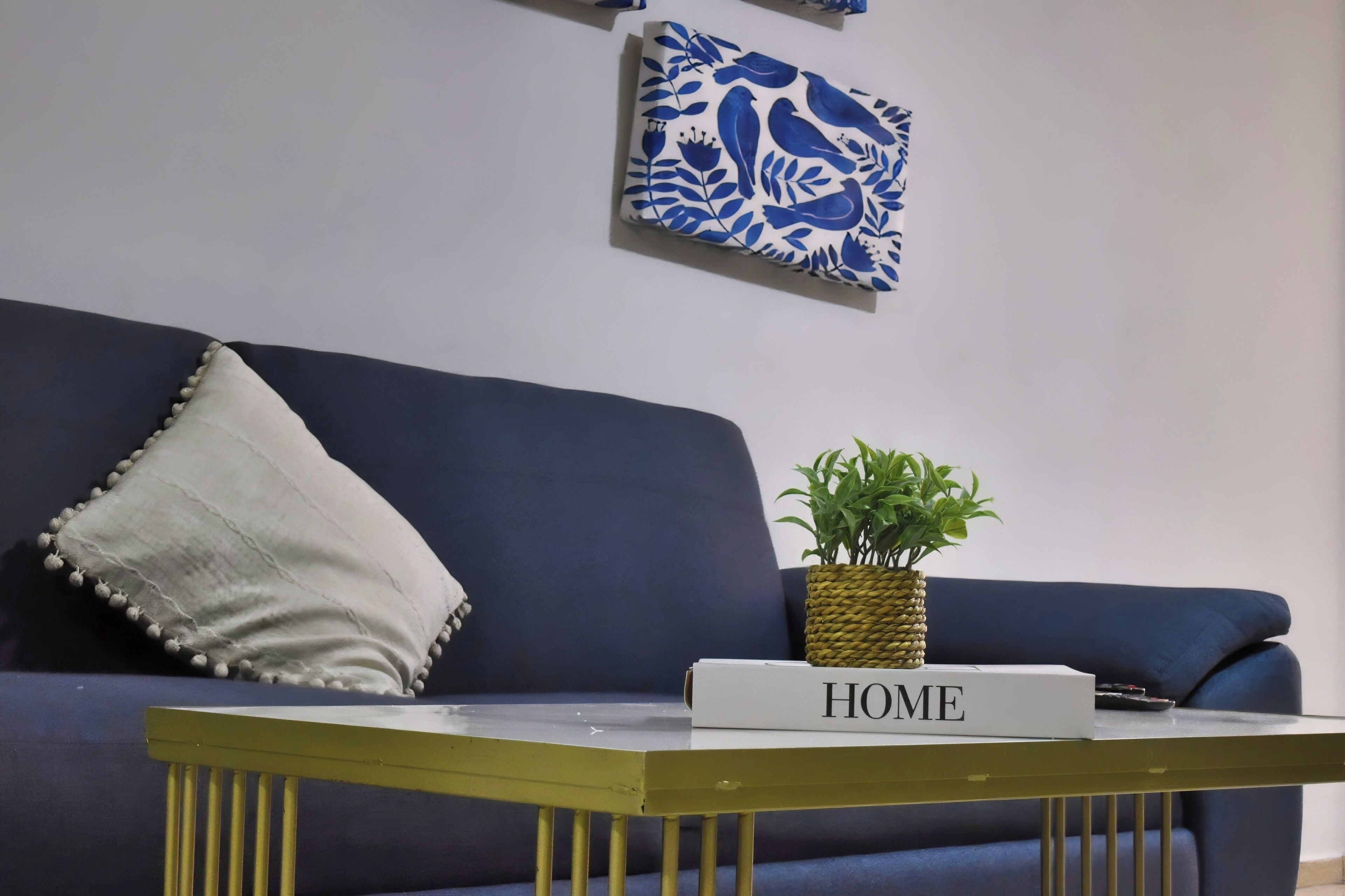 A blue sofa with a white pillow, a small potted plant, and a 'HOME' sign on a table in front of it.