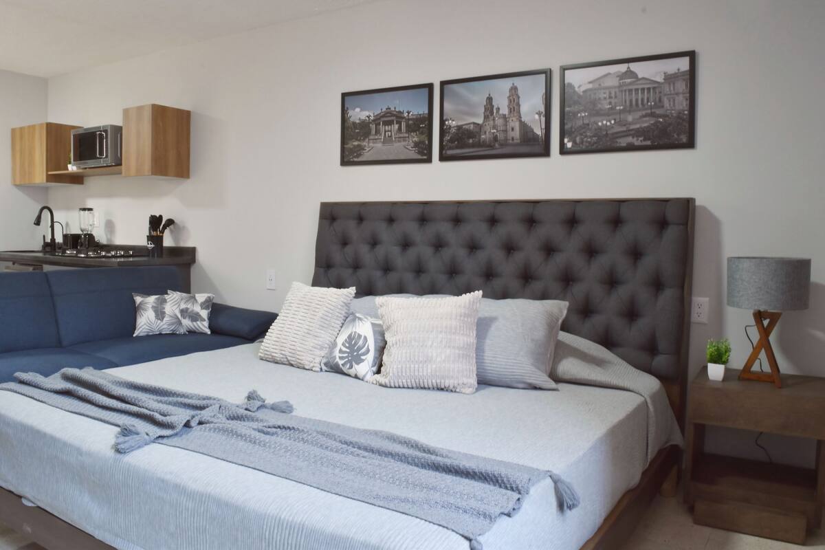 A bedroom with a large bed, a sofa, a kitchen area with cabinets, and three framed pictures on the wall.