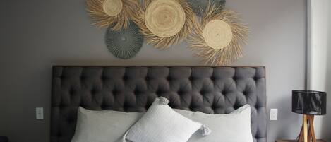 Quarto com cabeceira estofada, duas almofadas, mesa de cabeceira com abajur e obra de arte na parede.