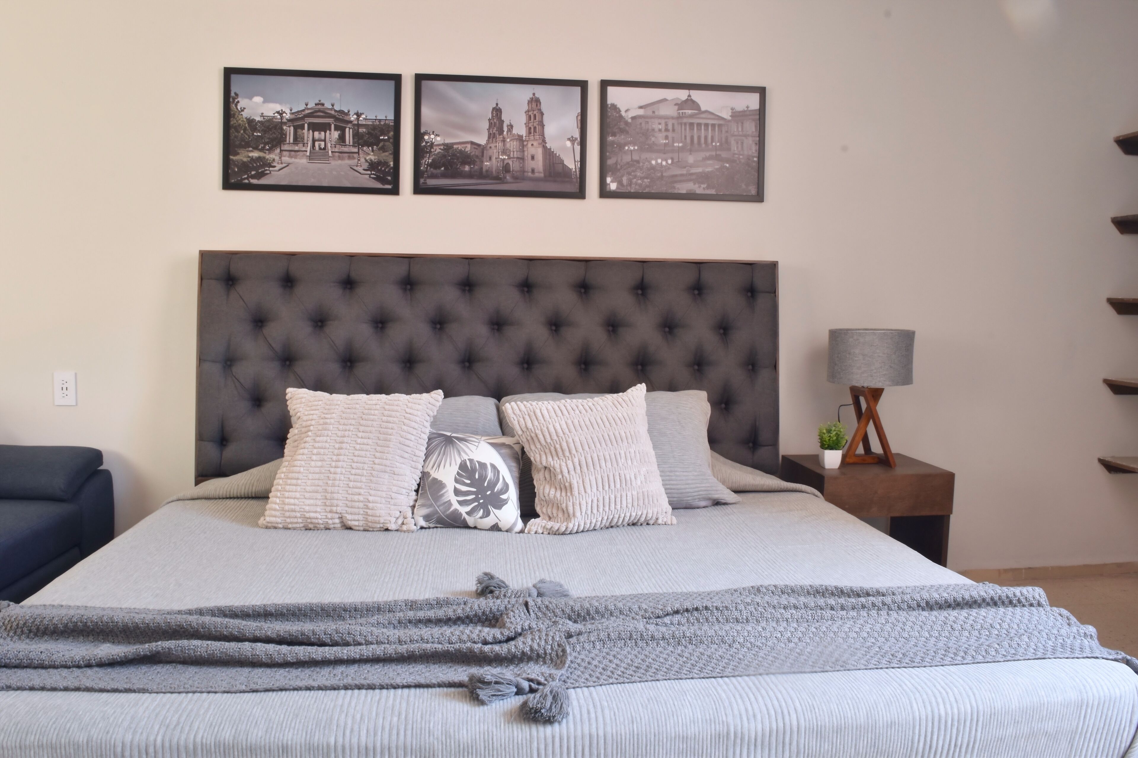 Un letto rifatto con cura, con una testiera grigia, numerosi cuscini e una coperta lavorata a maglia grigia. Sopra il letto, tre quadri appesi alla parete.
