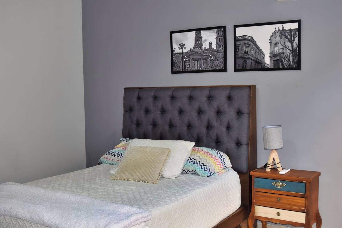 A bedroom with a tufted headboard, a bed with patterned pillows, a bedside table with a lamp, and two framed pictures on the wall.