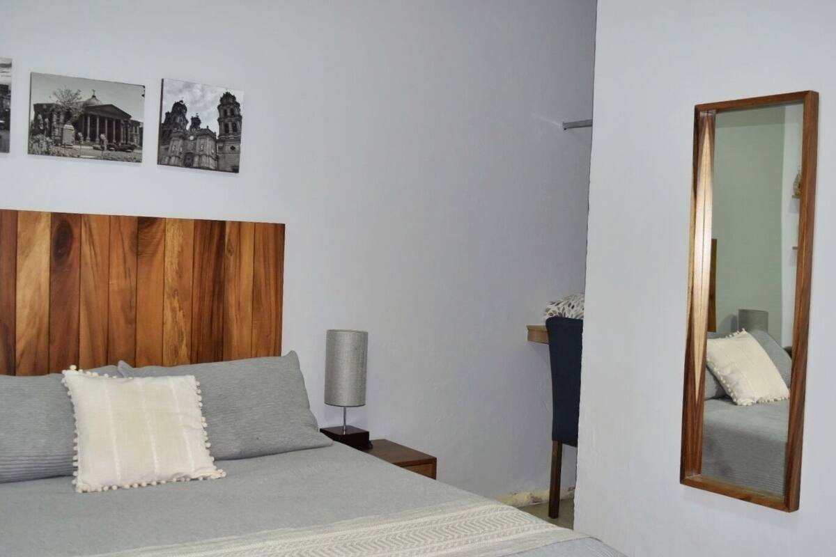 A bedroom with a wooden headboard, a bed with grey bedding, a nightstand with a lamp, and a mirror on the wall.