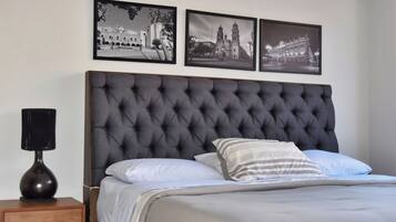 Une chambre à coucher avec une tête de lit capitonnée, un lit avec des oreillers blancs et à rayures, une table de chevet avec un vase noir, et trois photographies en noir et blanc accrochées au mur.