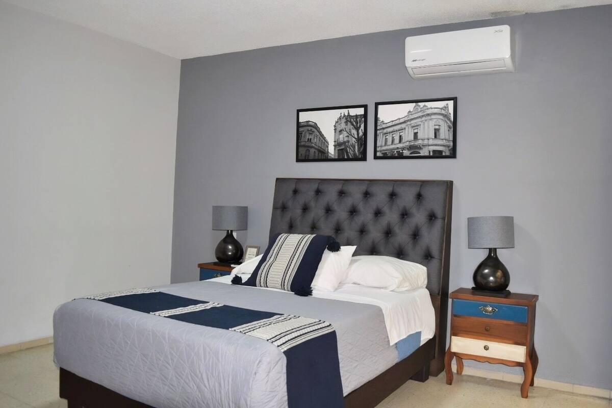 A bedroom with a large bed, two bedside tables, a wall-mounted air conditioner, and two framed pictures.