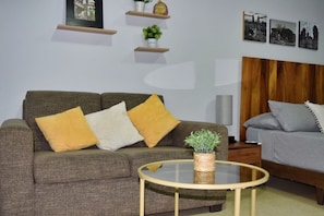 Sala de estar com sofá, mesa de centro redonda, mesa lateral, luminária e cama com cabeceira.
