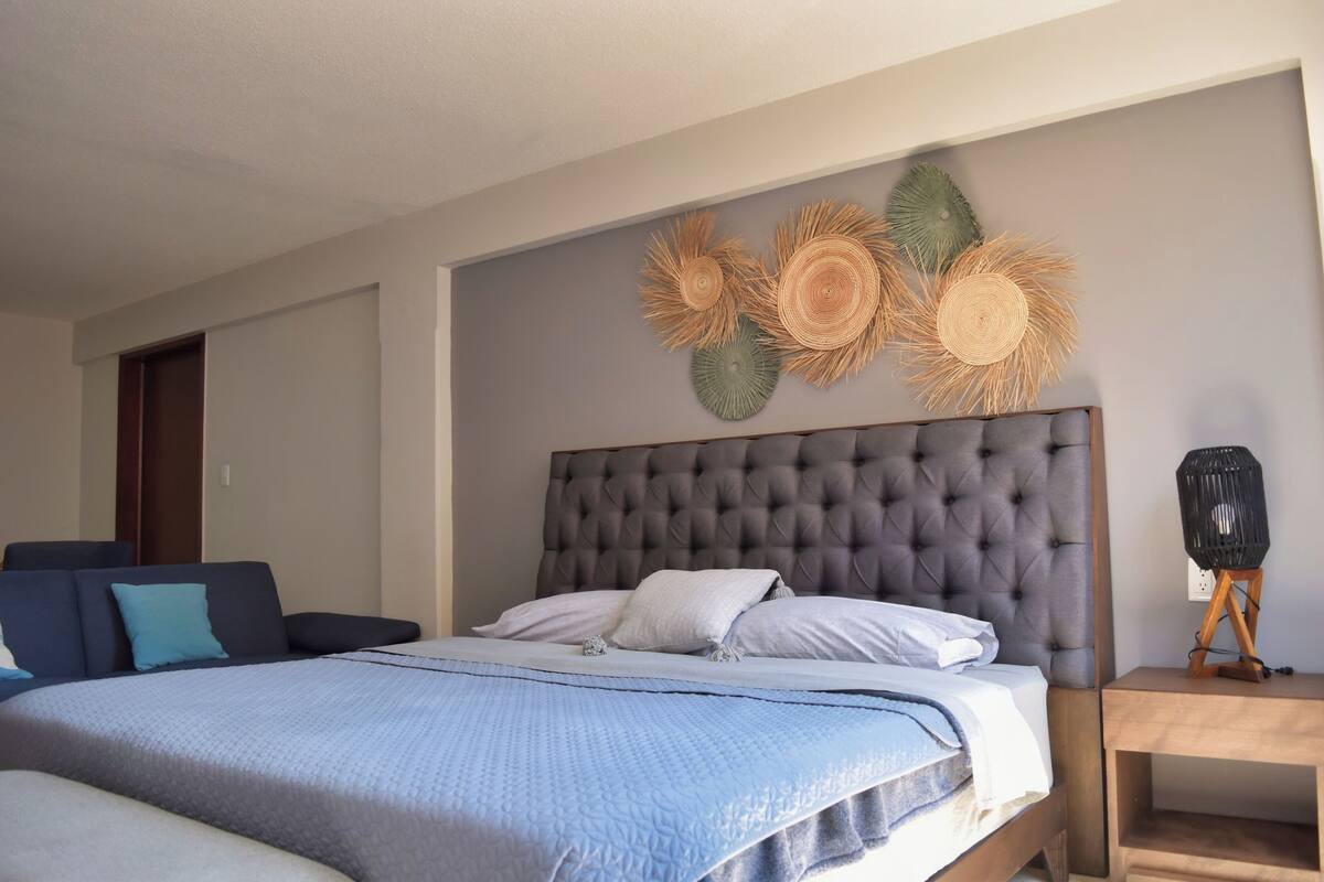 A bedroom with a tufted headboard, a bed with a blue quilt, a nightstand with a lamp, and wall art.
