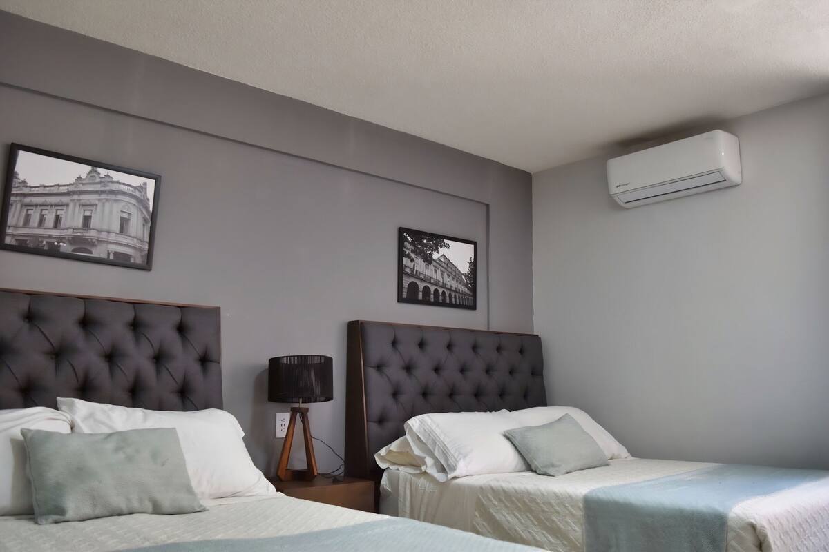A hotel room with two beds, a wall-mounted air conditioner, and two framed pictures.