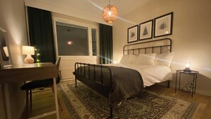 A bedroom with a bed, a desk, a chair, a nightstand, a lamp, a window with curtains, and framed pictures on the wall. - Magical and Cosy apartment with tree views from every window! (Rovaniemi)