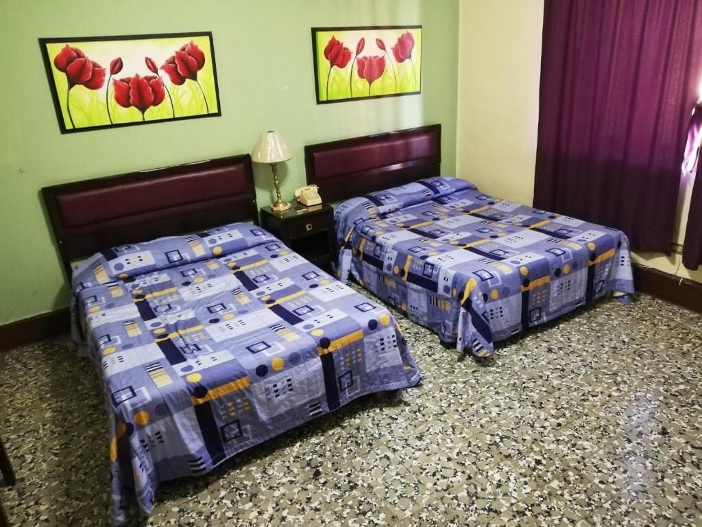 Ein Hotelzimmer mit zwei Betten, jedes mit einer gemusterten Tagesdecke, einem Nachttisch mit Lampe und zwei Bildern von roten Blumen an der Wand.