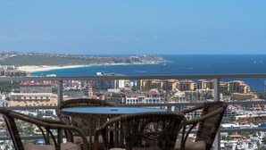 A balcony with wicker chairs and a table overlooking a coastal cityscape with buildings and a beach.