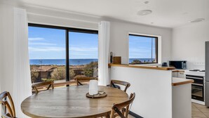 A modern kitchen with a view of the sea, a dining area with a wooden table and chairs, and a small island with a microwave. - Sapphire Tides - Stunning Views (Tura Beach)