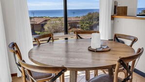A round wooden table with four chairs, a candle, and two glasses on a tray. A large window offers a view of the sea and nearby buildings. - Sapphire Tides - Stunning Views (Tura Beach)