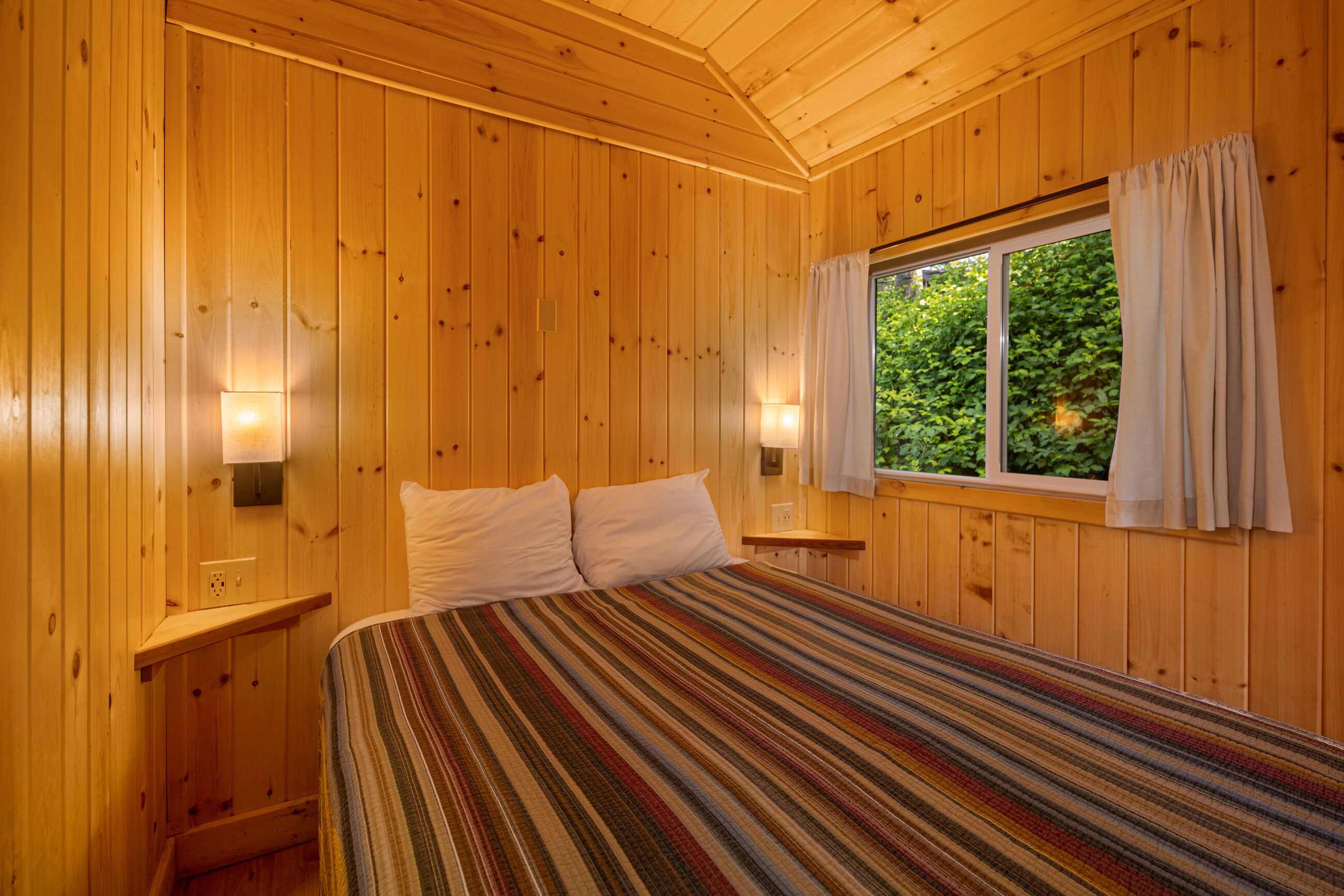 A wooden-paneled room with a bed, two bedside lamps, and a window with curtains.