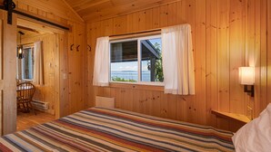 A wooden cabin room with a bed, a window with curtains, a chair, and a lamp. - 2BR Beach Cabin on prime west-facing beach! (Eastsound)