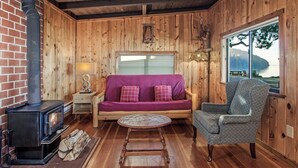 A cozy cabin interior with a wood-burning stove, a purple sofa, a round wooden table, and a view of a lake through a large window. - 2BR Beach Cabin on prime west-facing beach! (Eastsound)