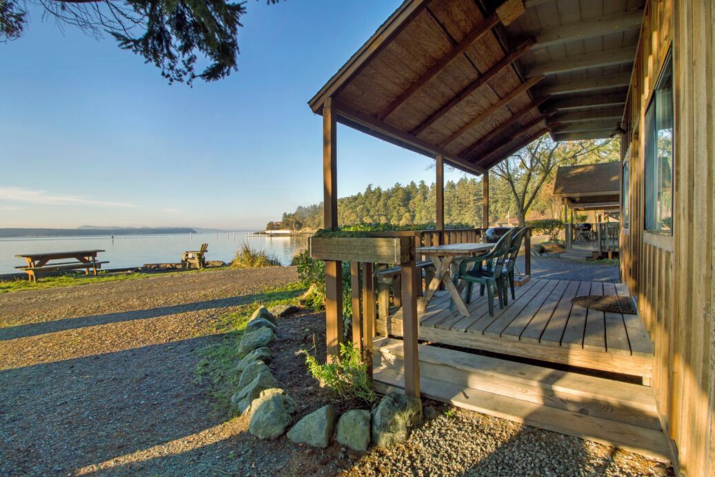 Una terraza de madera con mobiliario exterior, con vista a un lago y un área boscosa.