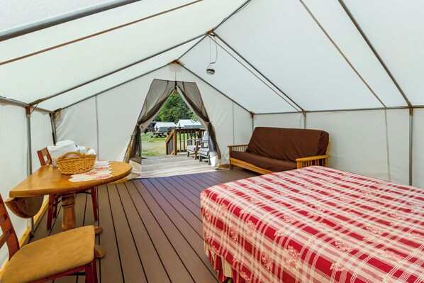 A tent with a bed, a table, and a chair. There is a view of the outside through a window.