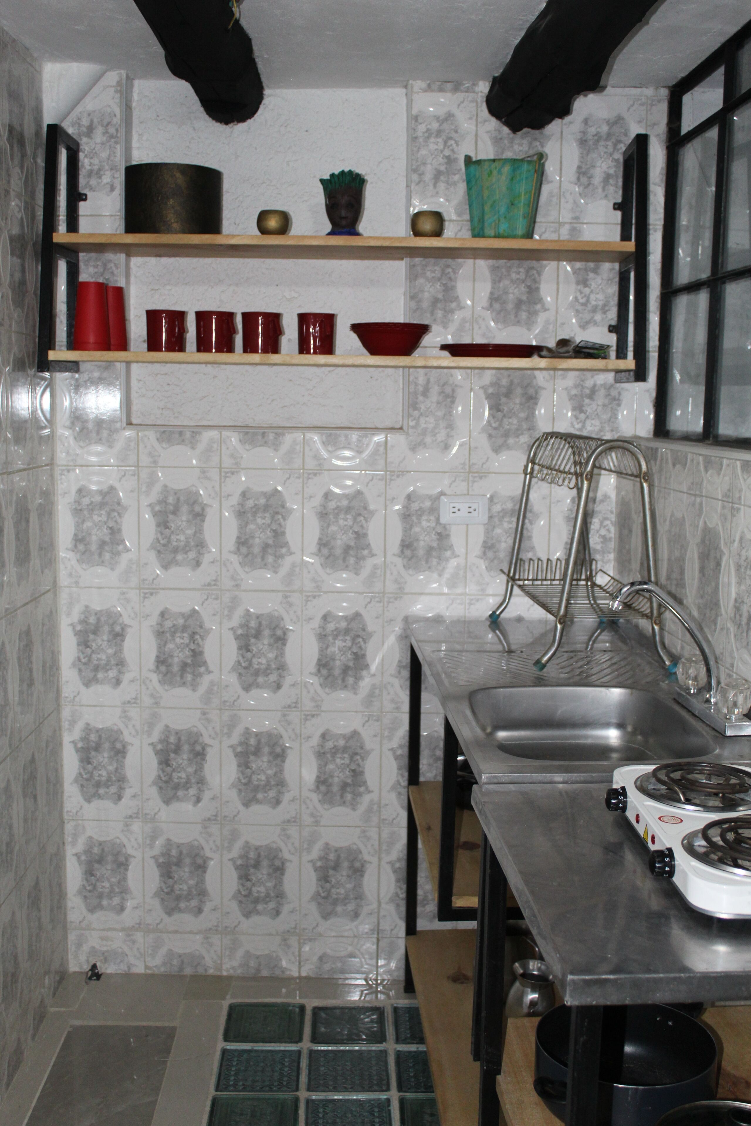 A compact kitchen with a sink, stove, and shelves with red cups and a green pot.