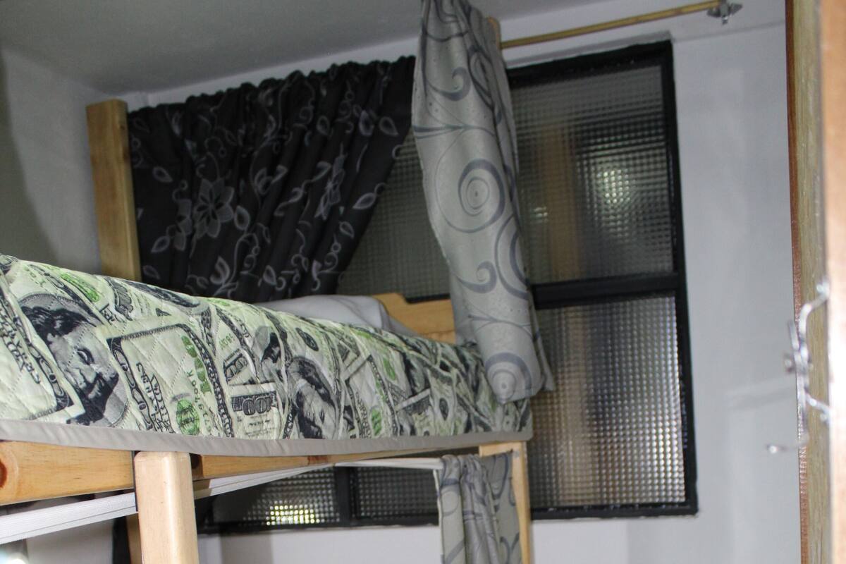 Ein Stockbett mit gemustertem Bettbezug, eine Holzleiter, ein kleiner Nachttisch und ein Fenster mit einem Metallgitter.